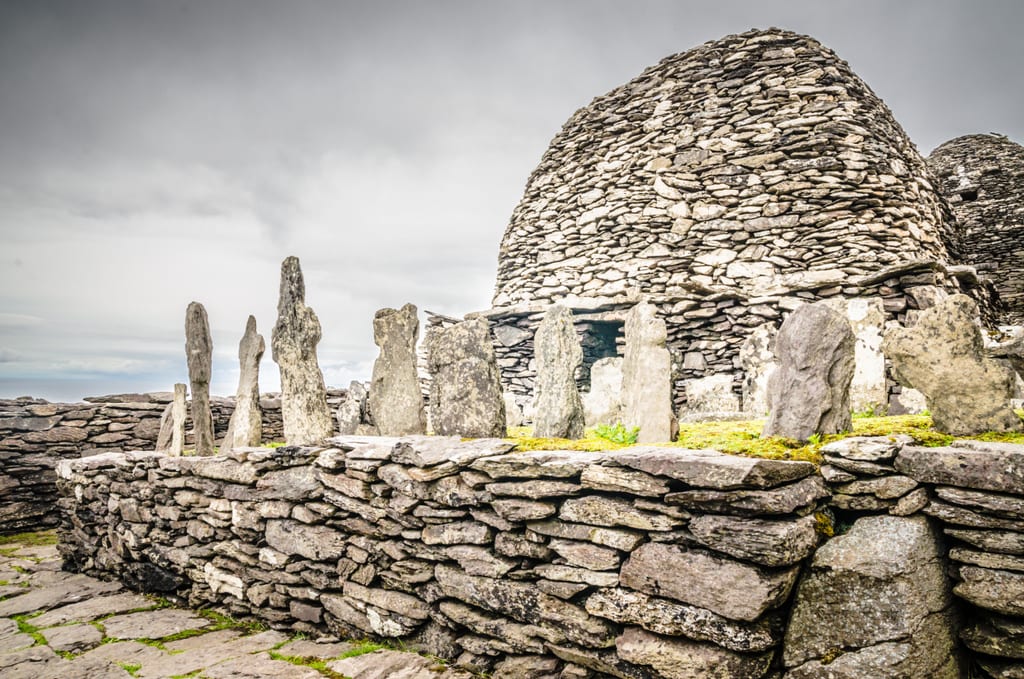 Rows of plain stone gravestones in front of one of the rocky beehive-shaped huts.