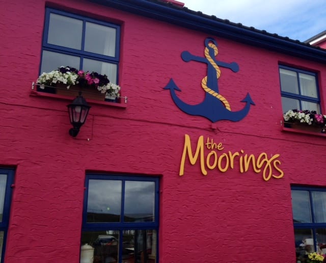 A bright red guesthouse with windows, a navy blue anchor, and text that reads "The Moorings" in yellow.