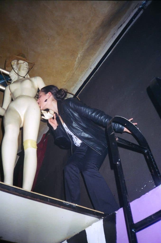 Kate standing on a tall platform kissing a mannequin on the butt.