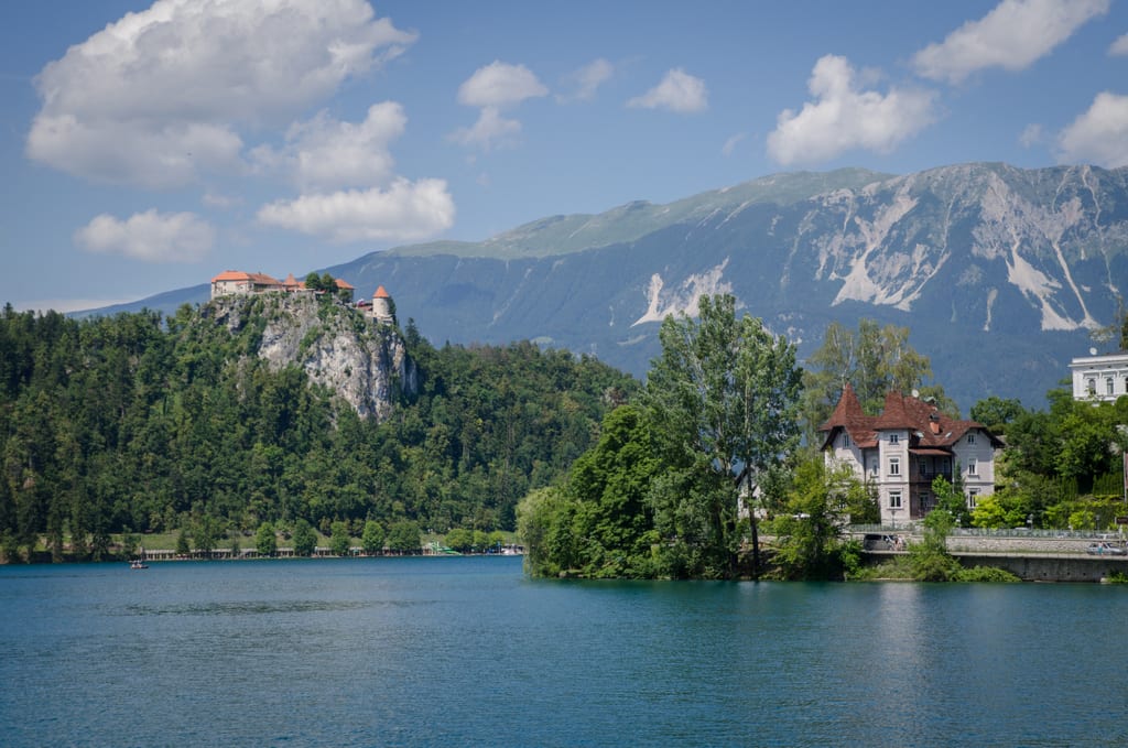 Lake Bled: a soft blue lake with mountains in the background. On the left is a castle perched on top of a cliff right next to the lake; on the right is a smaller white building in the foreground.