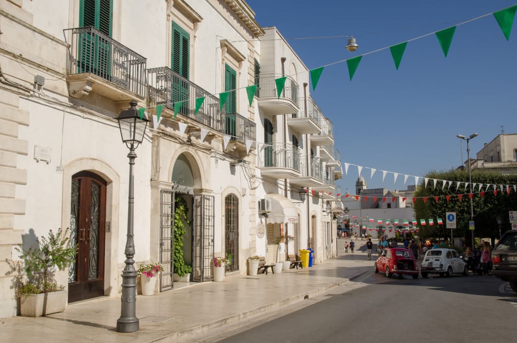 A street in Italy with white buildings and green flags hanging between the buildings.