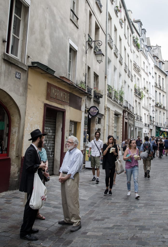 A Paris street with white buildings, people walking down the street and having conversations, including an orthodox Jewish man in a black hat.