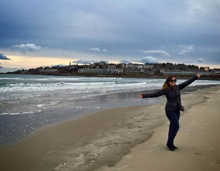 Kate standing on the beach in York Maine on a gray winter day, her arms up in the air like jazz hands.
