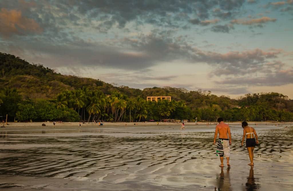 Two people walking on a gray beach underneath a blue sky with pink and gray clouds in Samara, Costa Rica.