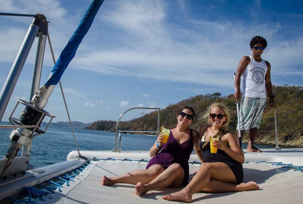 Kate and Alex sitting on the net of a Catamaran with a crew member in the background, in front of bright blue ocean.