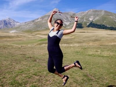 Kate wears overalls and jumps in the air in front of mountains.