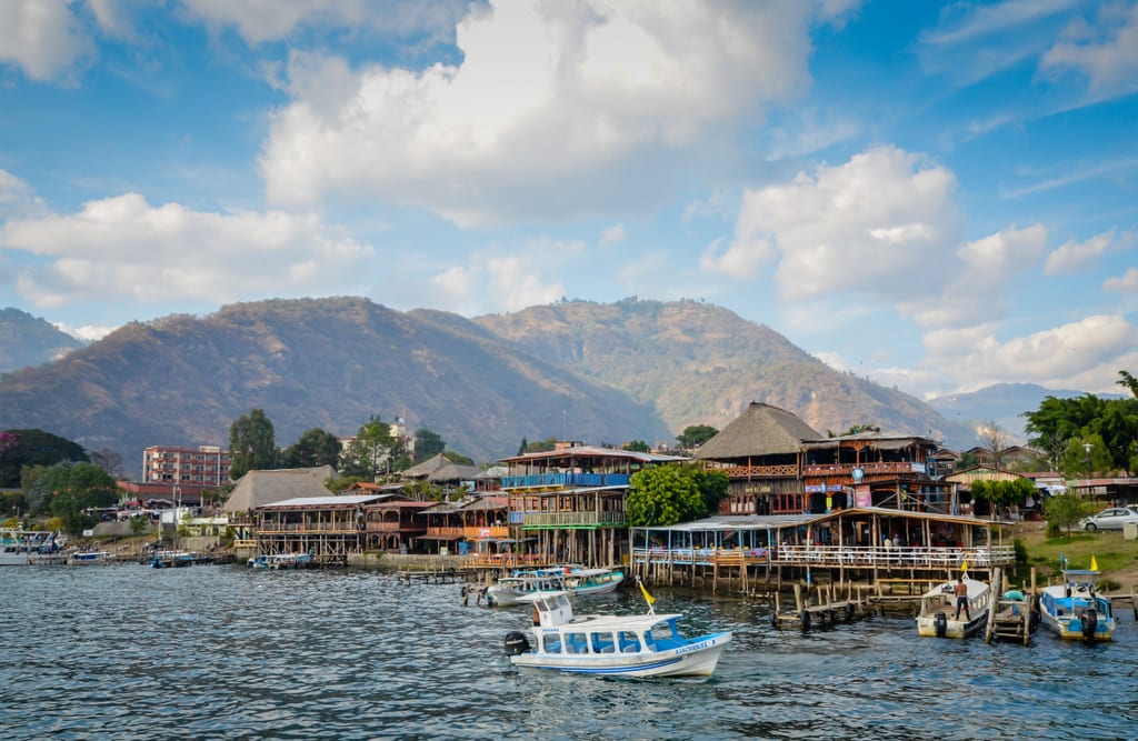 Ramshackle wooden buildings built on stilts on the shore of a lake, mountains rising in the background.