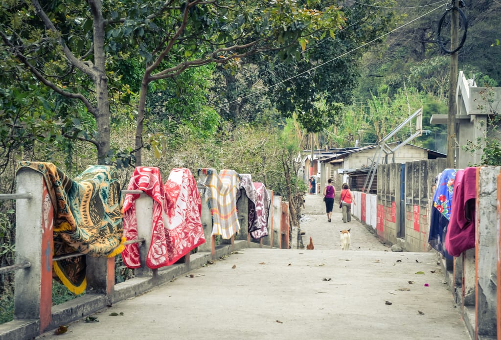 Women walking down a street past a bridge covered with traditional blankets.