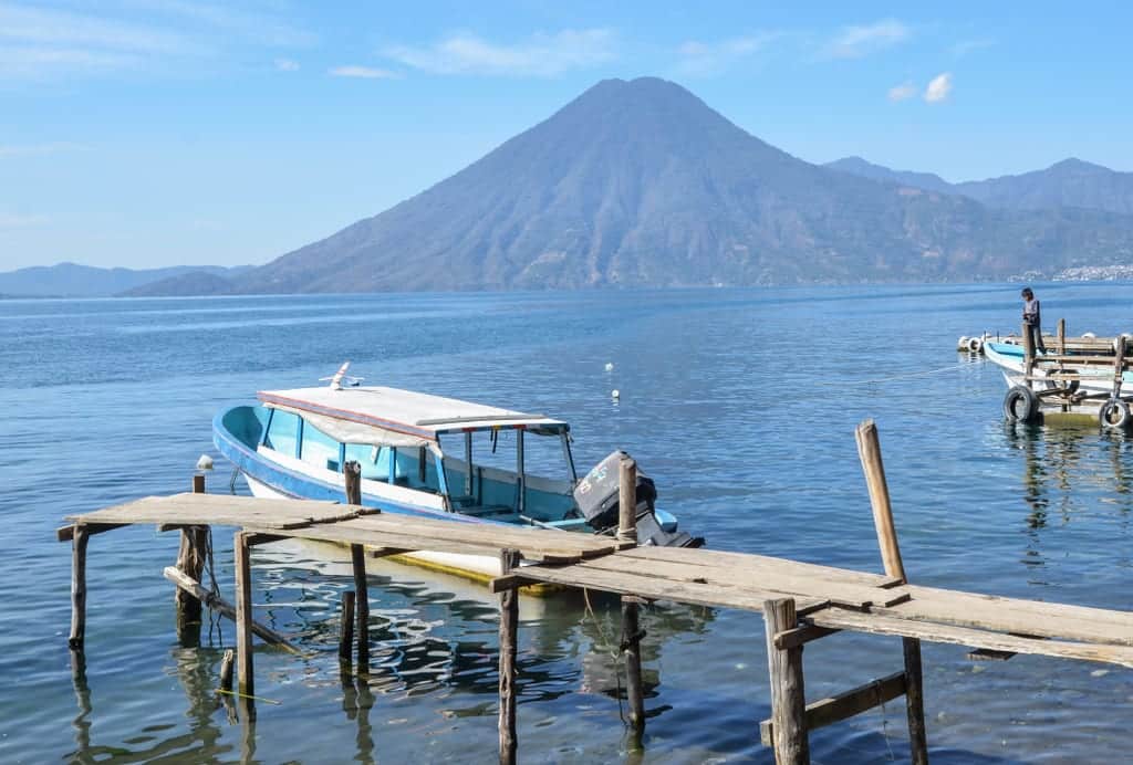A single white boat parked on a ramshackle wooden dock in front of a volcano on the lake.