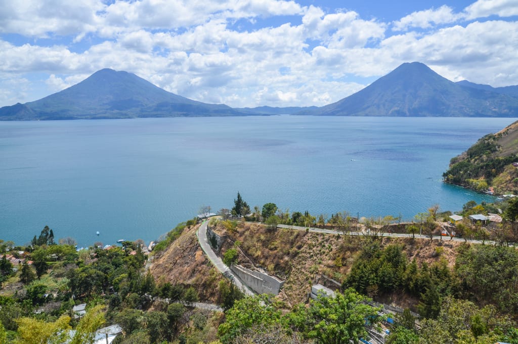 A road winding around steep mountain passes in front of the bright blue lake with its two pointy volcanoes.