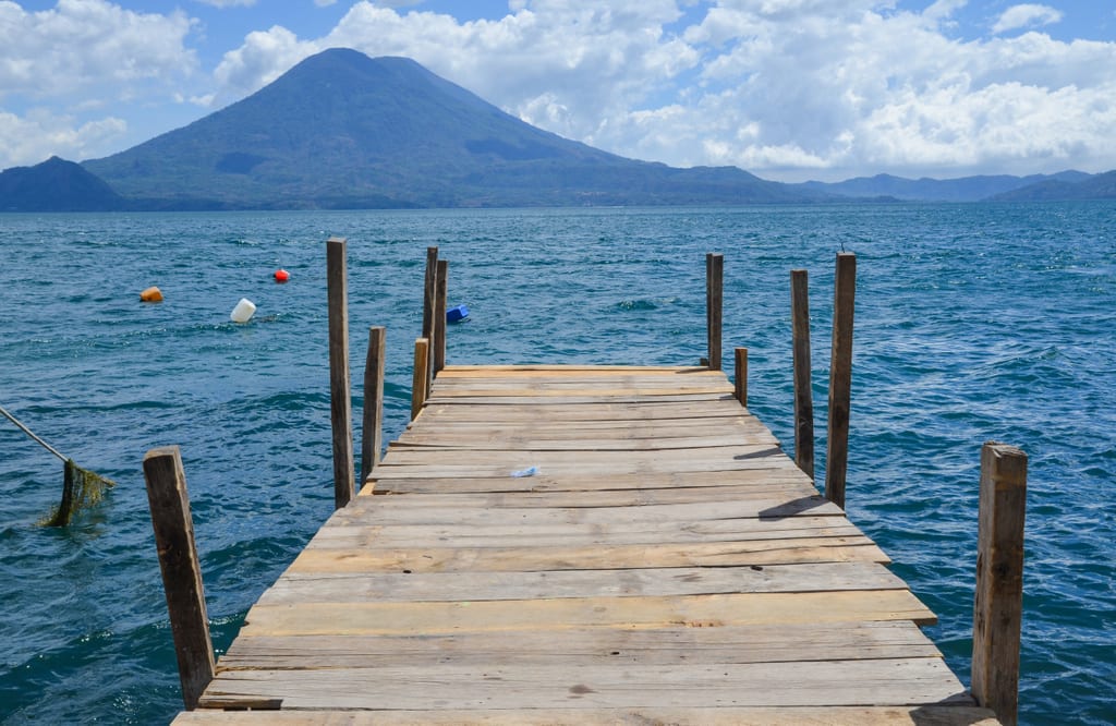 A wooden dock leading out to a bright blue lake, a pointy volcano in the background.