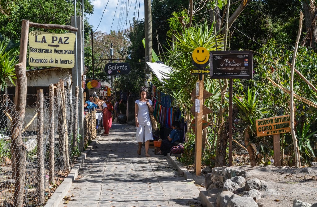 A woman walking down a street surrounded by lush forest.