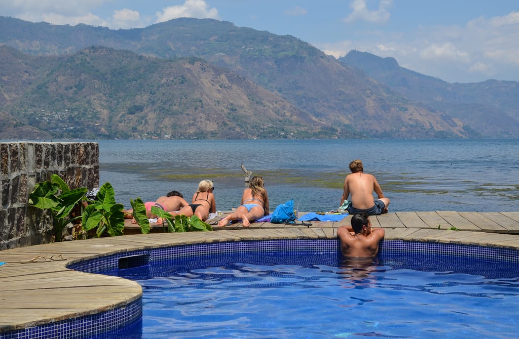 A few backpackers tanning in the sun next to a pool in front of the lake.