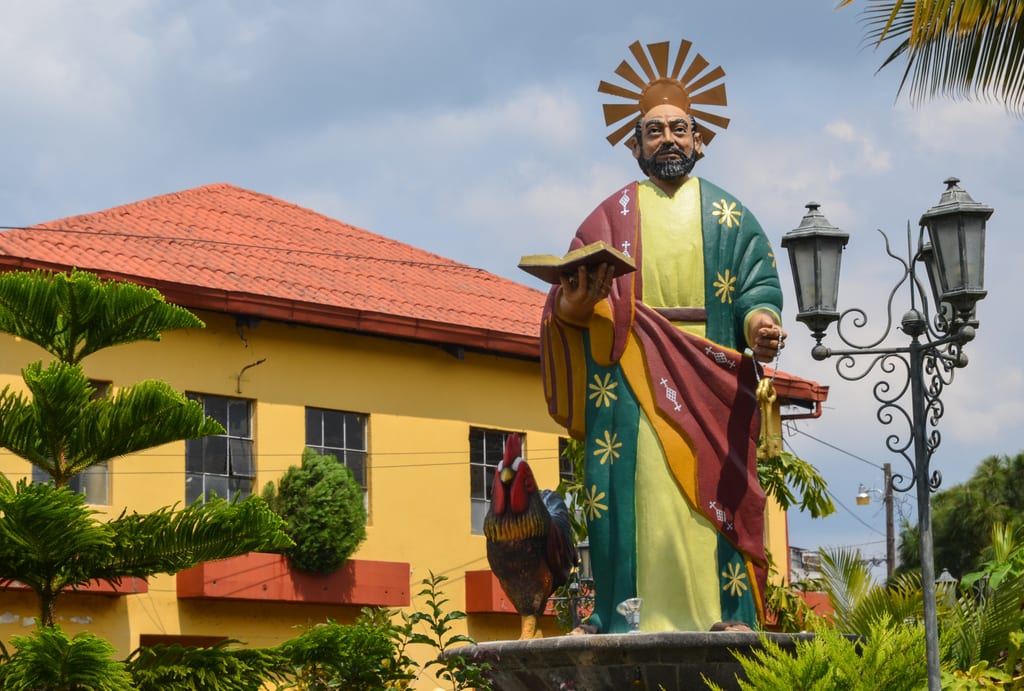 A large statue of a saint-like man with a golden sunburst behind his head, walking next to a rooster.