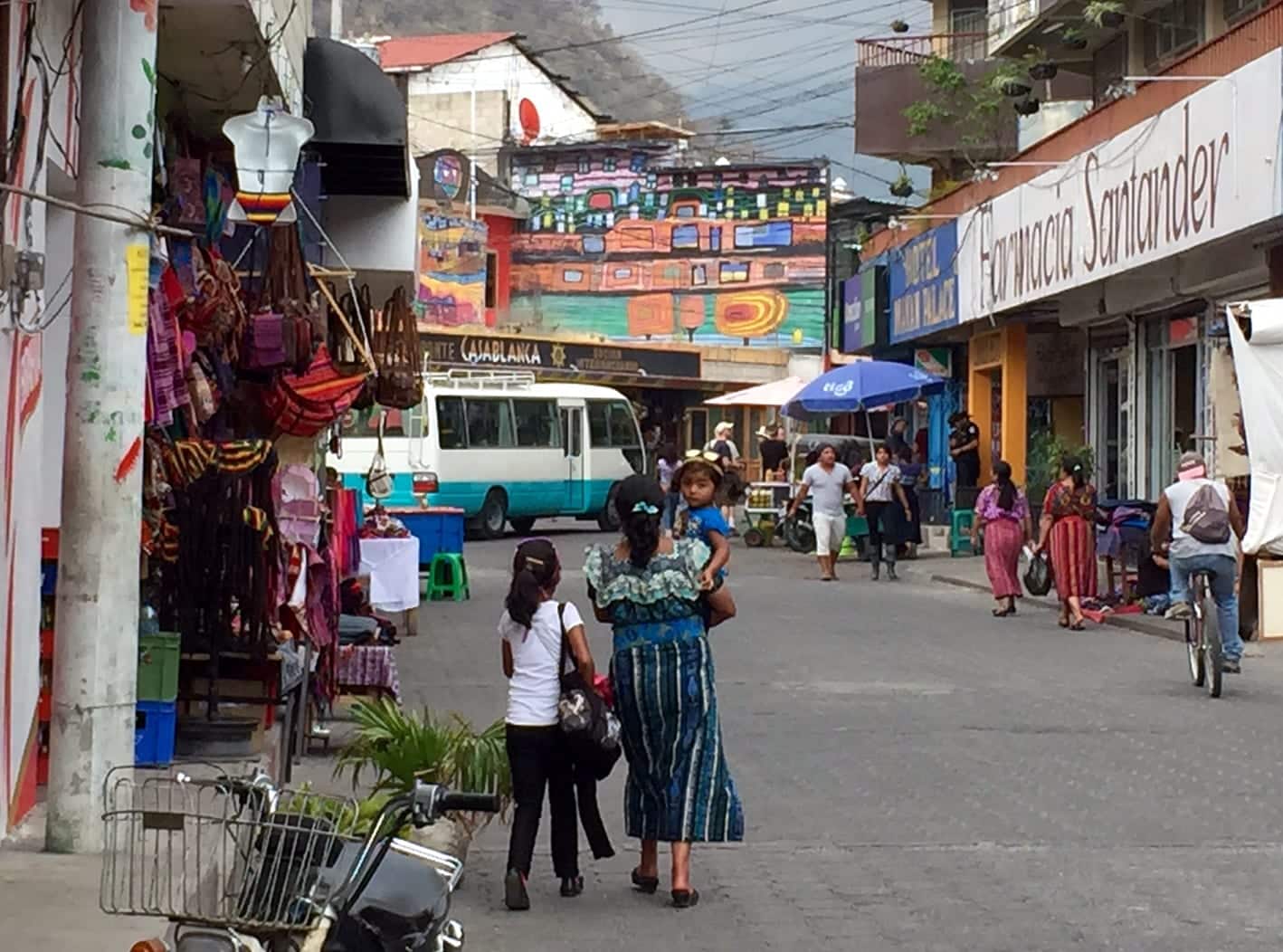 An Indigenous woman in a frilly blue blouse and striped woven skirt holding her daughter and walking down the street.