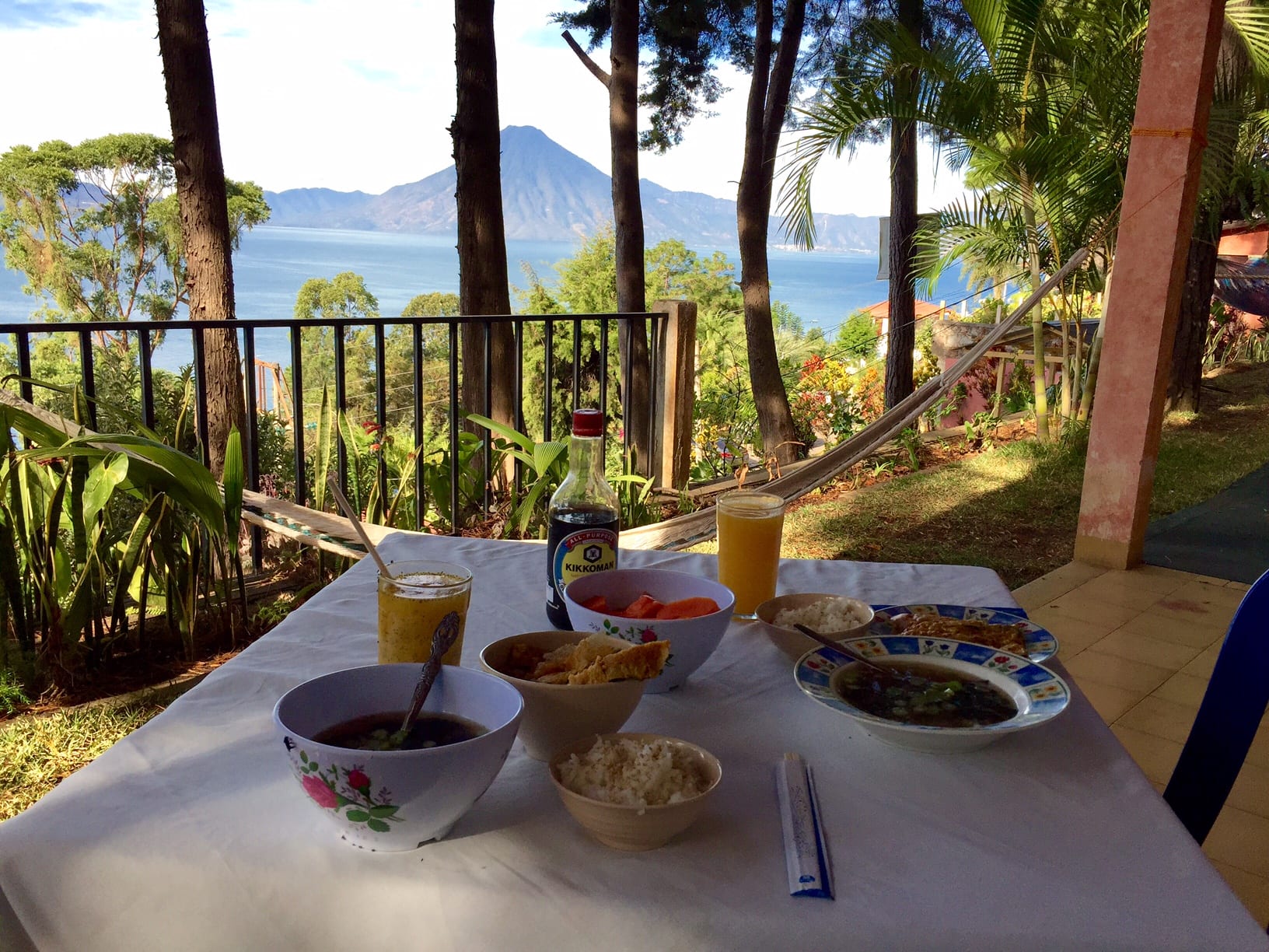 A table set for a Japanese breakfast of miso soup, rise, and fruit, with a hammock in the background and the lake with volcanoes on it beyond that.