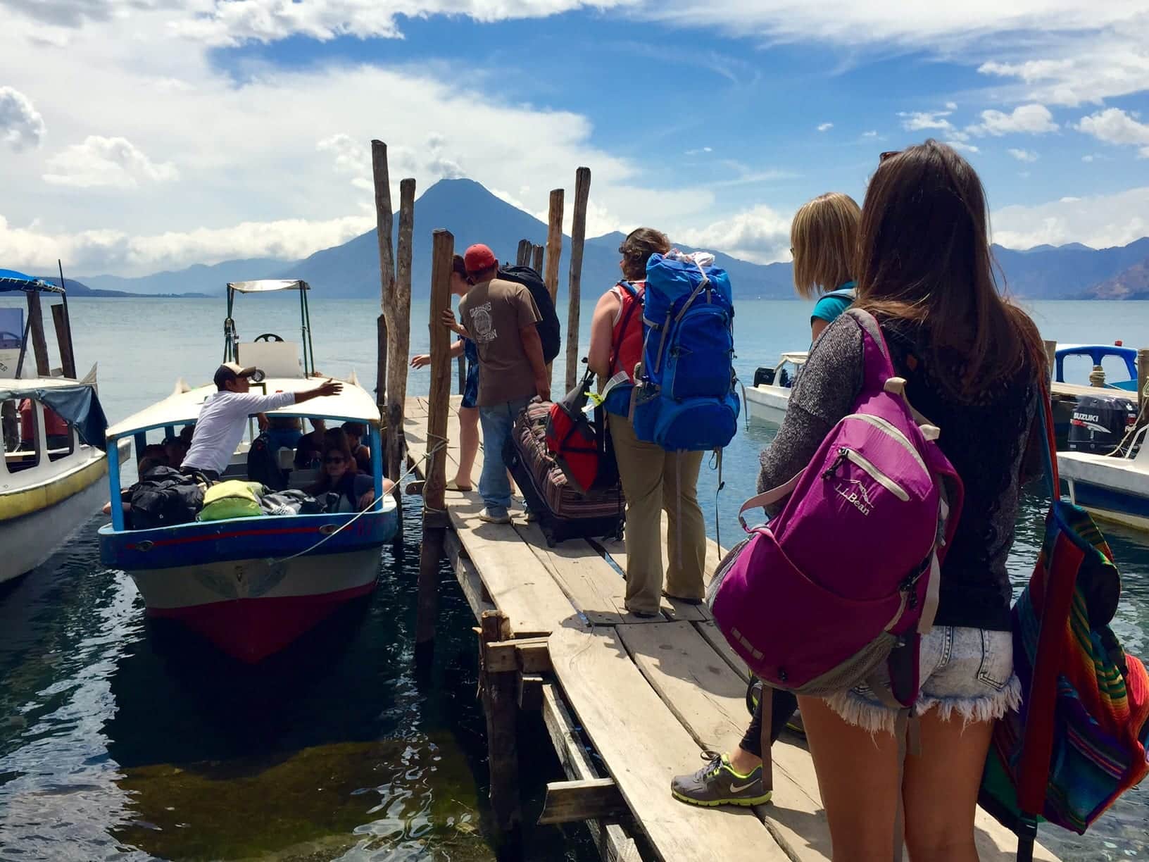 A group of travelers standing on a narrow dock as men pile their backpacks onto a small boat.