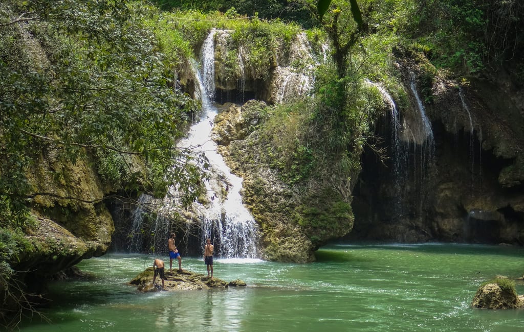Three young men standing near a waterfall emptying into a bright green river in Semuc Champey.