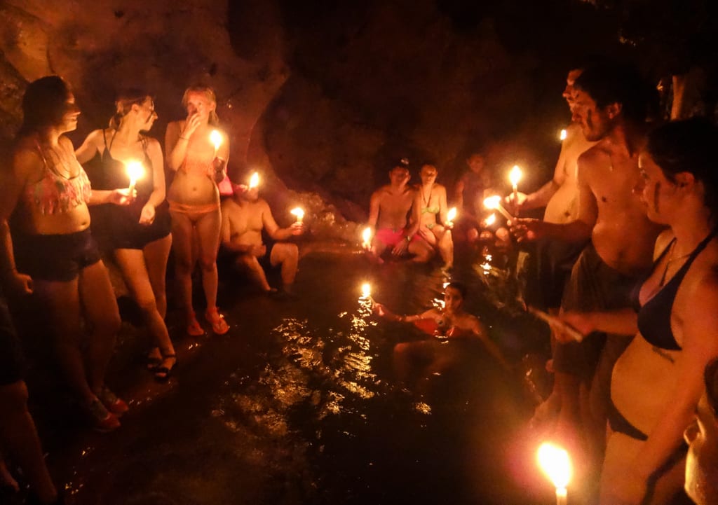 A group of people standing around a pool in the semuc champey caves holding candles. One girl is in the water, floating while wearing a life vest.