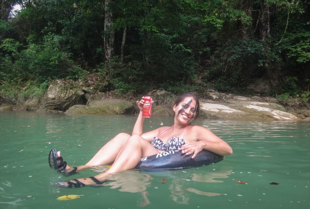 Kate in a tube in the river, holding up a can of beer and smiling, still with the black paint on her face.