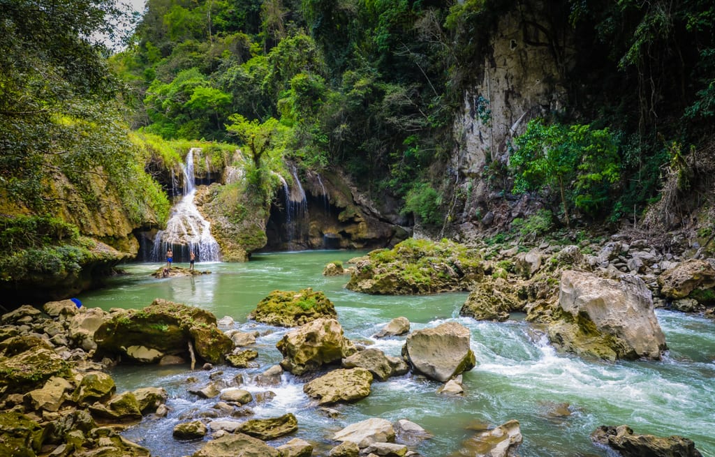 A small waterfall at Semuc Champey leading to a rocky green stream, two boys standing in front of the waterfall.