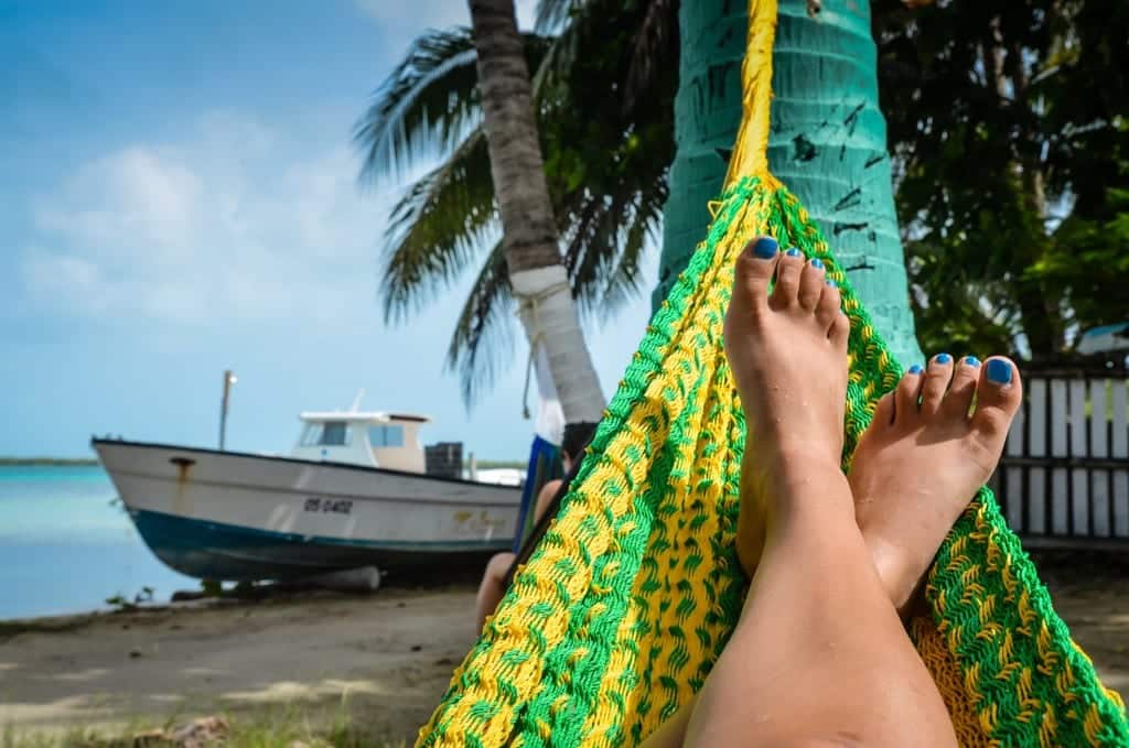 Feet in a green and yellow hammock with a boat on the beach in the background