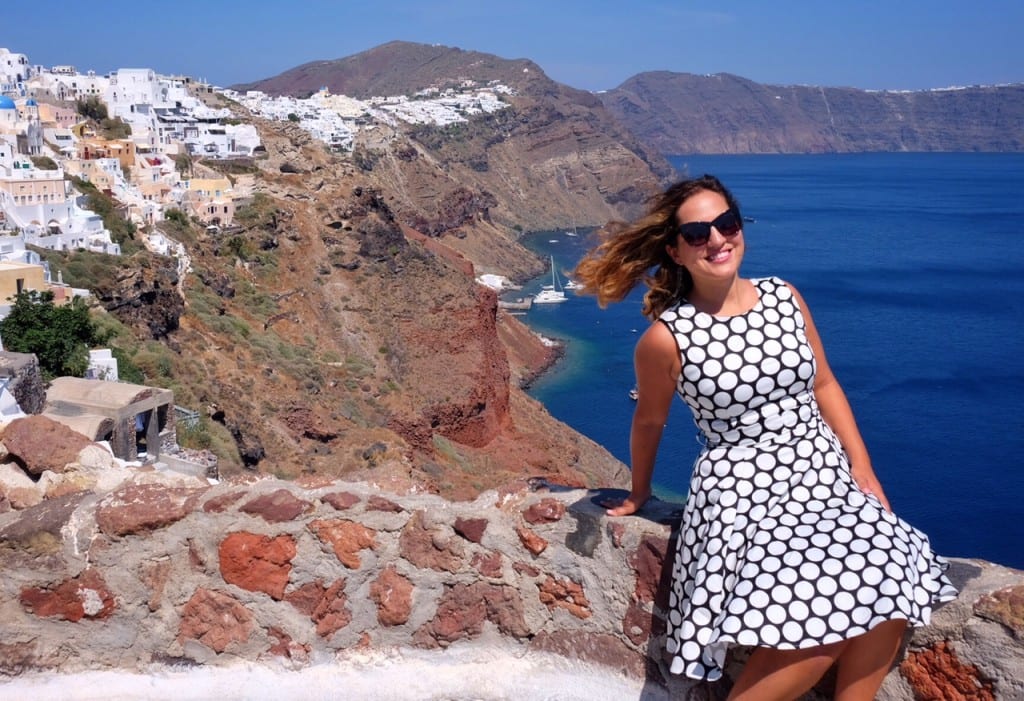 Kate wearing a black and white polka dot dress, standing in front of a cliff topped with white villages in Santorini.