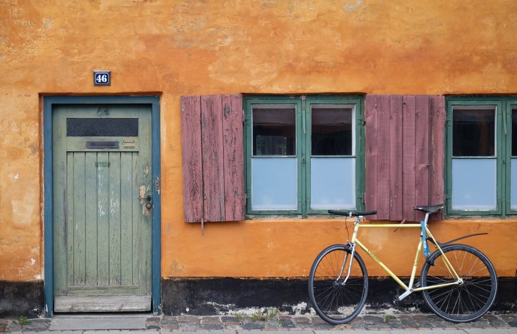 A old-fashioned orange house in Copenhagen with raspberry shutters and a green door. A bike is parked in front.