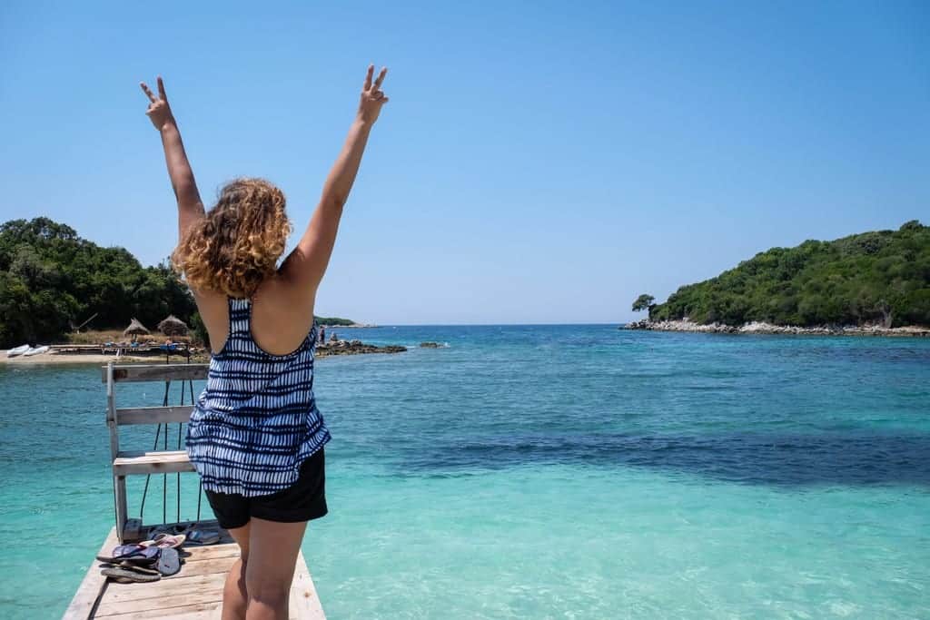 Kate faces away from the camera with her arms in the air in front of the turquoise water of Ksamil, Albania