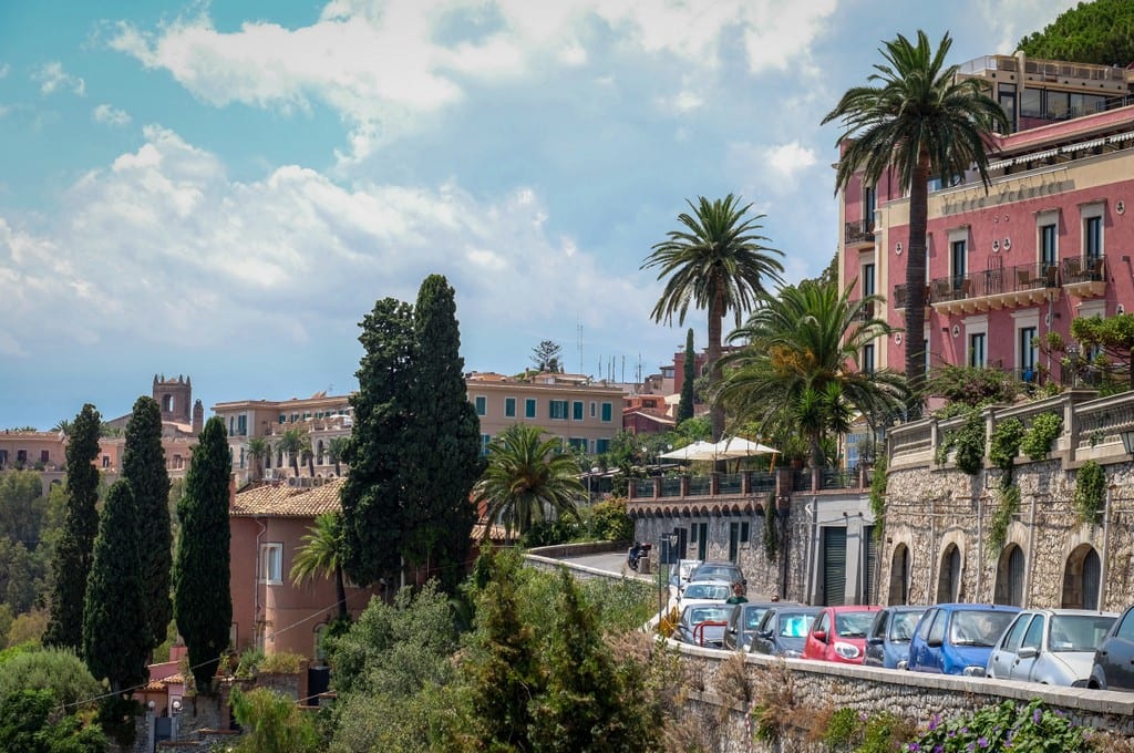 Bright pink buildings, tall cypress trees, and winding roads on the edge of a hill in Sicily.