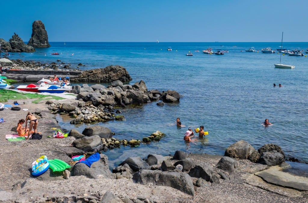 People relaxing on beach towels on a rocky outcropping in Aci Trezza, Sicily, leading to the bright blue sea, filled with boulders.