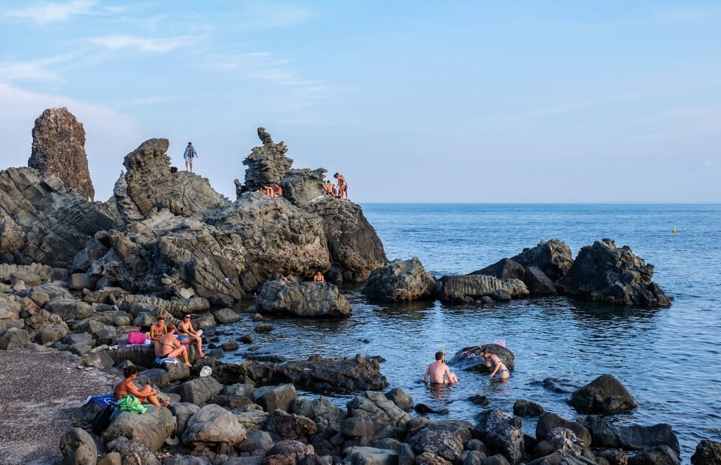 People hanging out on the rocky coastline in the blue water of Aci Trezza, Sicily