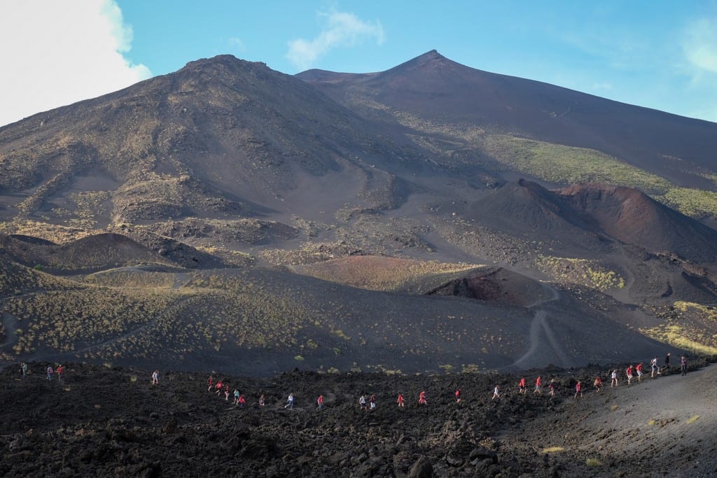A line of people in orange vests walking along a dark ridge of a tall volcano,
