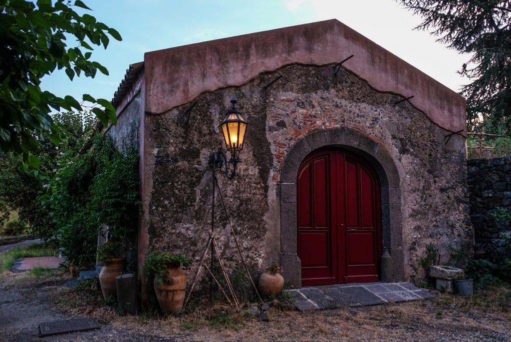 A pink building in Italy with a lantern, a big wooden door, surrounded by overgrown plants.