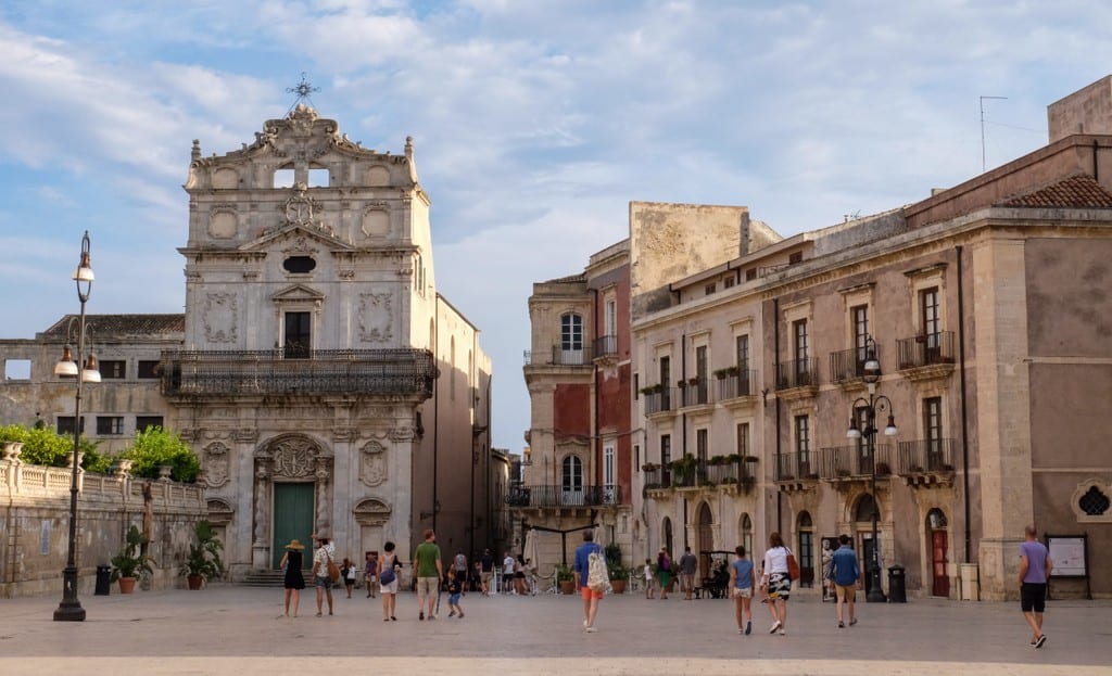 A piazza in Siracusa, Sicily, filled with stone churches and buildings. Lots of kids running around.