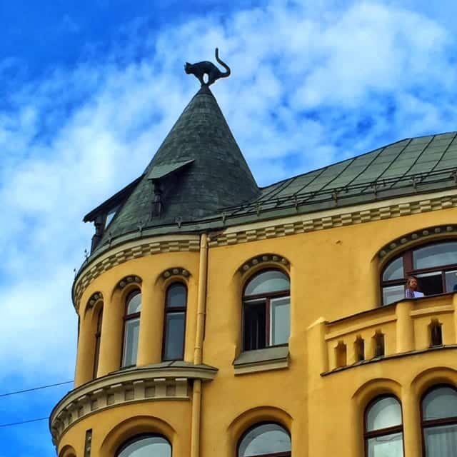 A yellow crenellated building in Riga. Its turret is topped with a black metal cat.