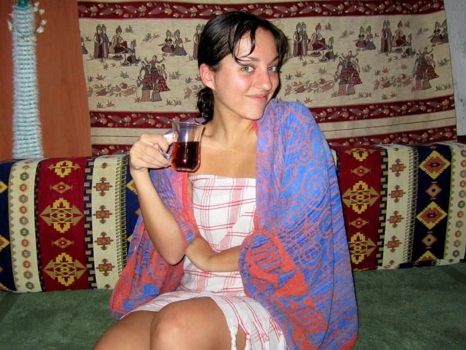 Kate sits on a patterned sofa while drinking a cup of tea, wrapped in towels with a mischievous smile on her face after finishing the Turkish hamam.