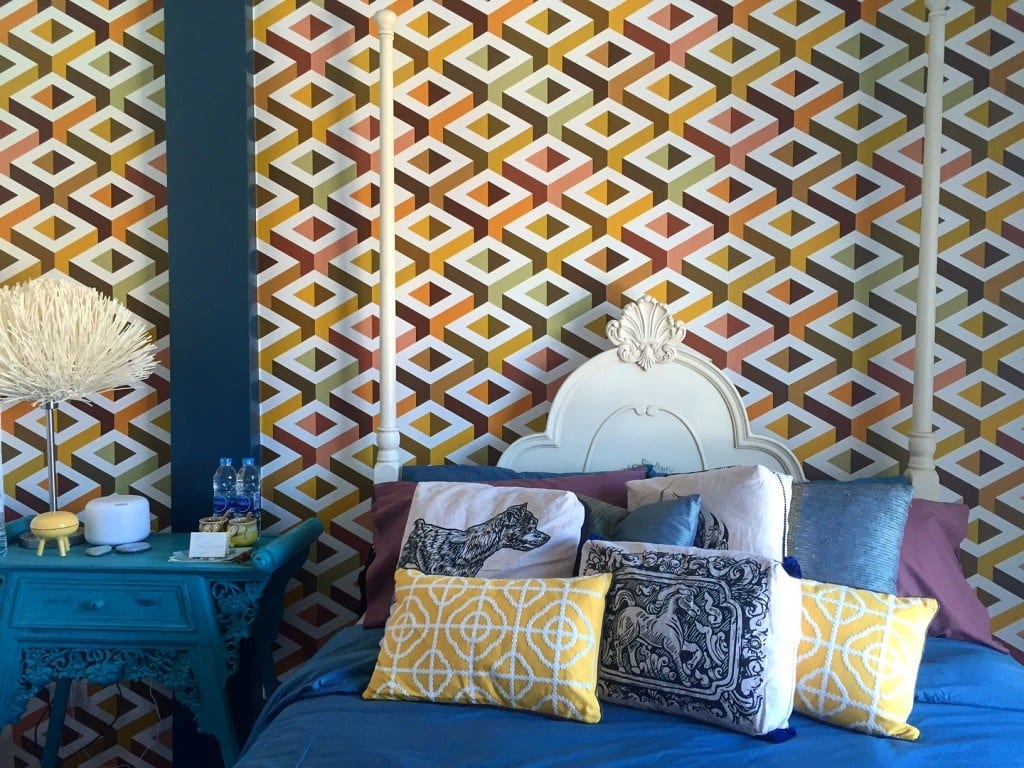 A collection of yellow and blue bellows on top of a blue bed with a curvy headboard in front of yellow diamond-patterned wallpaper.