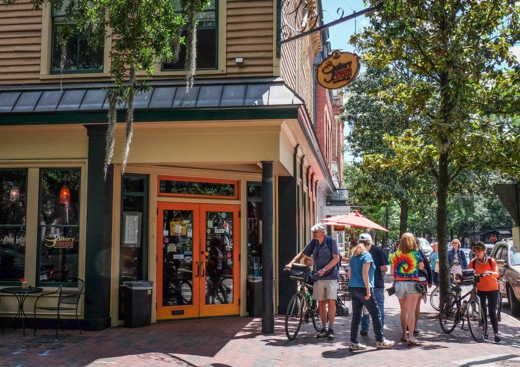 People walking and biking on the sidewalk in front of historic buildings in Savannah.