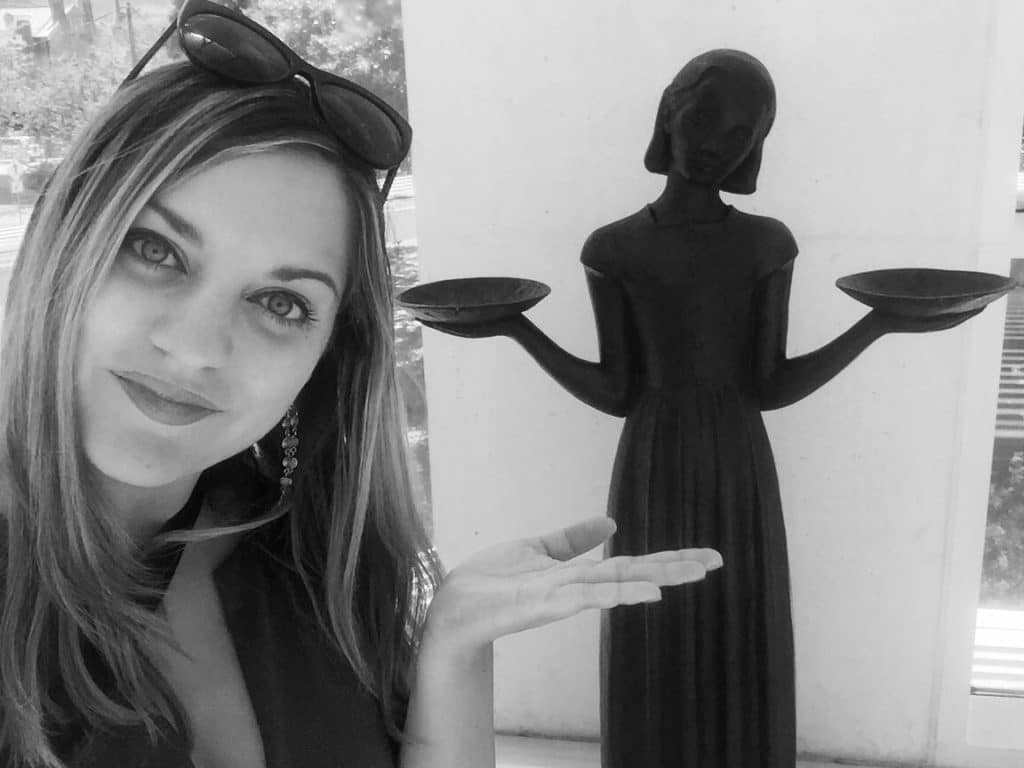 A selfie of Kate and a metal statue of a woman with her head cocked, holding a plate in each hand.