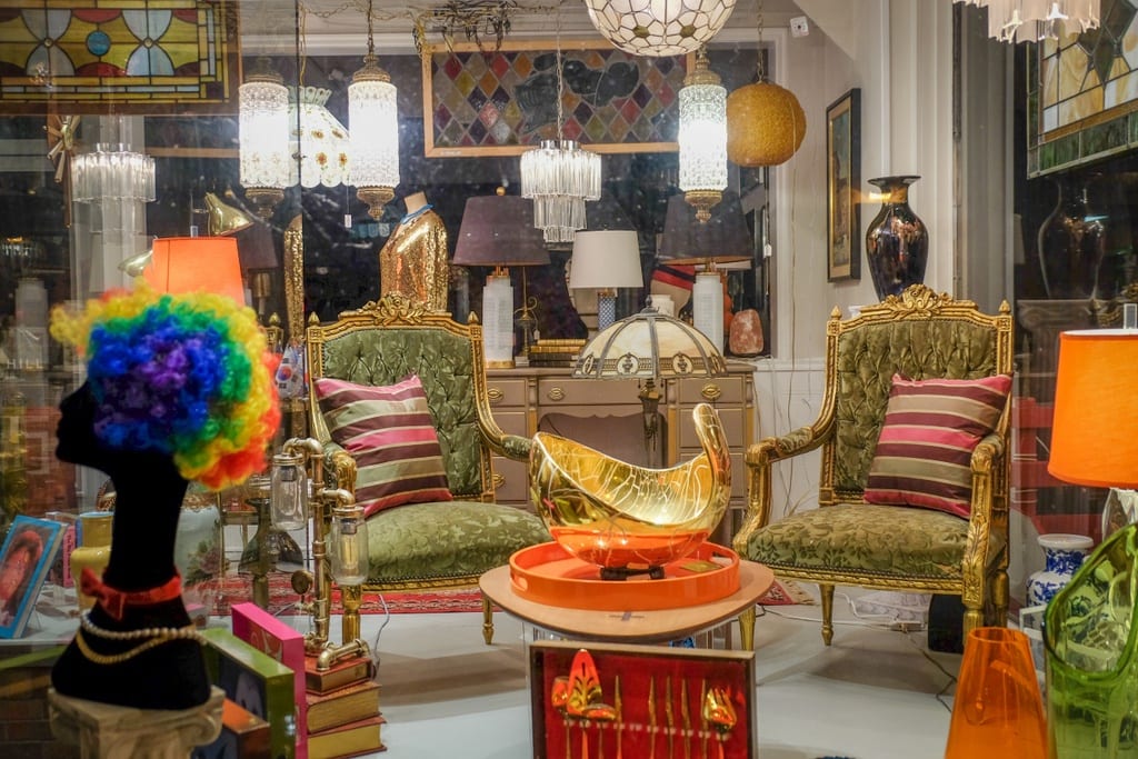 An antique store filled with green velvet and gold chairs, lamps, sculptures, a curly rainbow wig, and more.