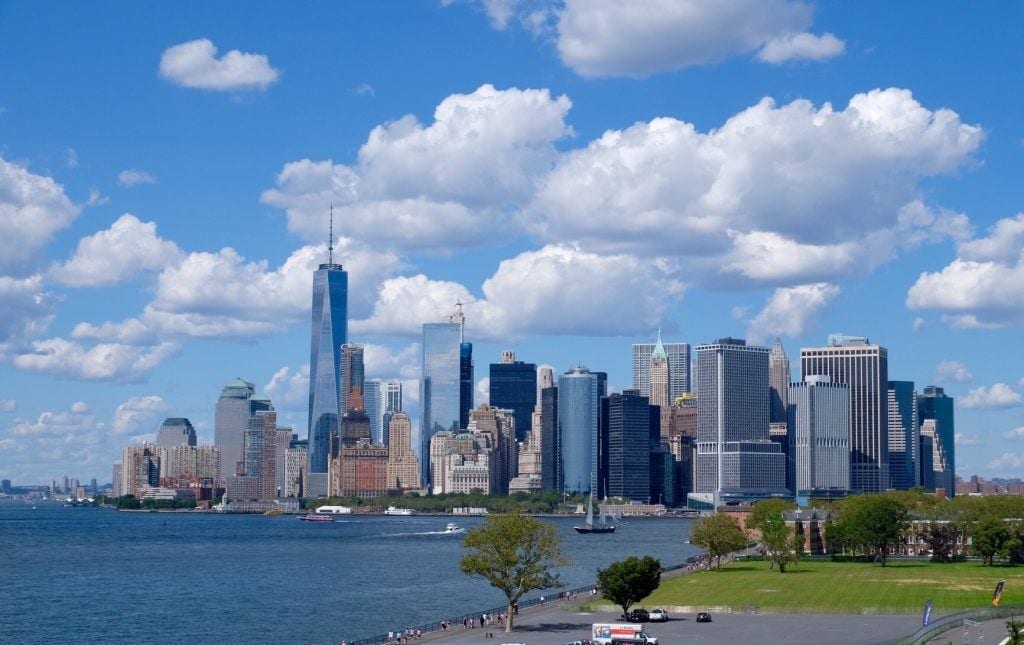 visit ny in november The Manhattan skyline underneath a partly cloudy sky, buildings rising up in shades of blue and gray.