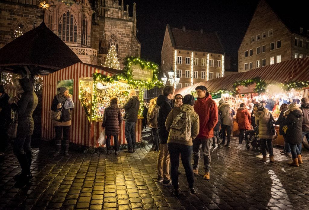 People gathered on a cobblestone square at Nuremberg Christmas Markets, surrounded by booths selling Christmas crafts.