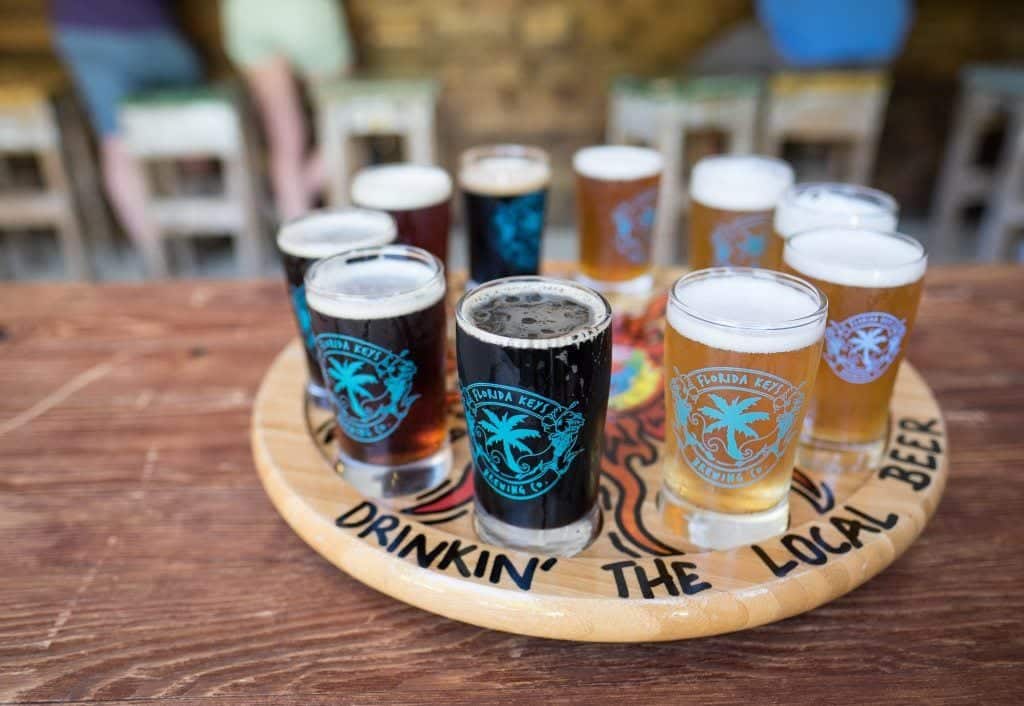 A round wooden sampler platter topped with several small glasses of beer.