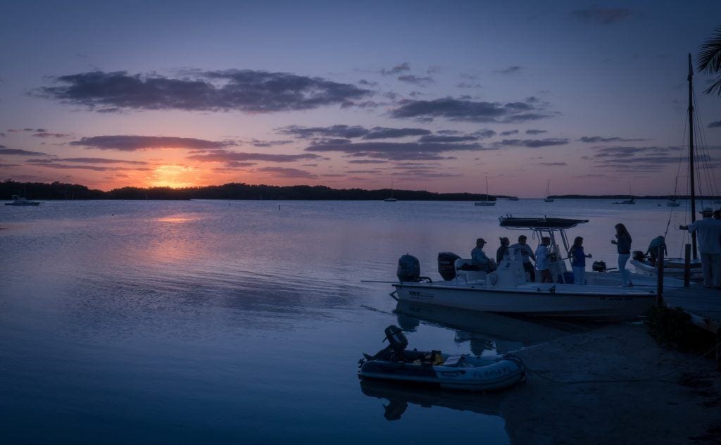 A white speedboat filled with people cruising along the water, the last yellow light of sun sinking into the blue sea.