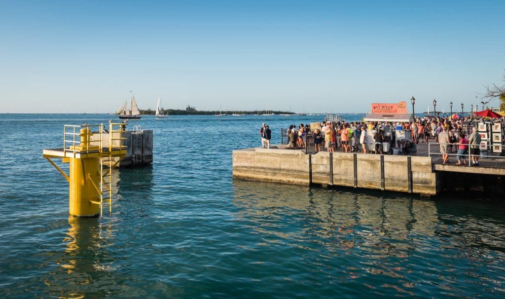 A pier jutting out into the bright blue water in Key West, lots of people standing on it to view the sunset.