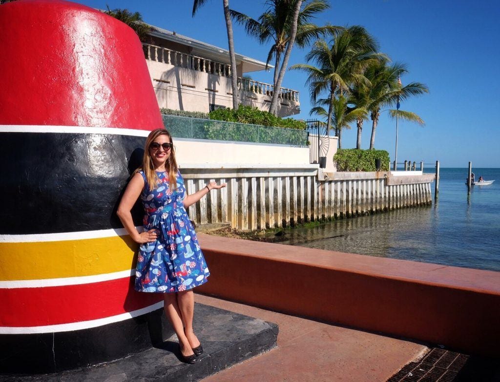 Kate wears a bright blue knee-length 50s housewife style dress and has long blonde straight hair, standing in front of the red, black, and yellow stone monument marking the southernmost point in the continental United States.