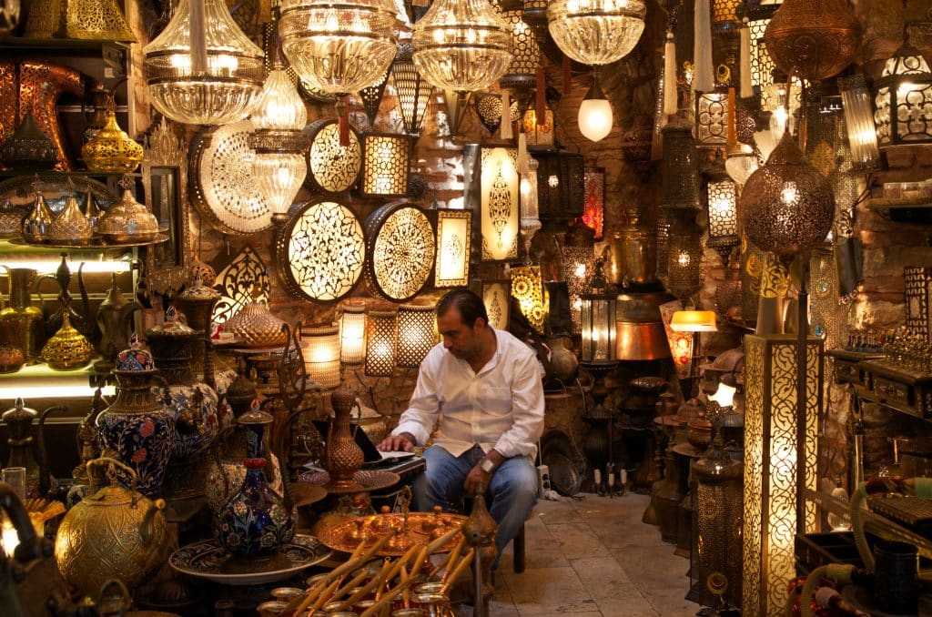 A man sitting in a shop in the Grand Bazaar surrounded by brightly lit lamps.
