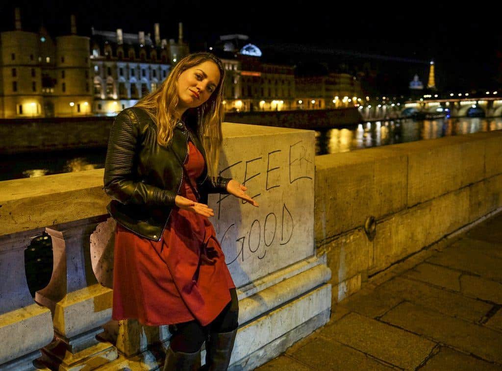 Kate in a red dress and leather jacket on a bridge in Paris emblazoned with "FEEL GOOD" in Graffiti.