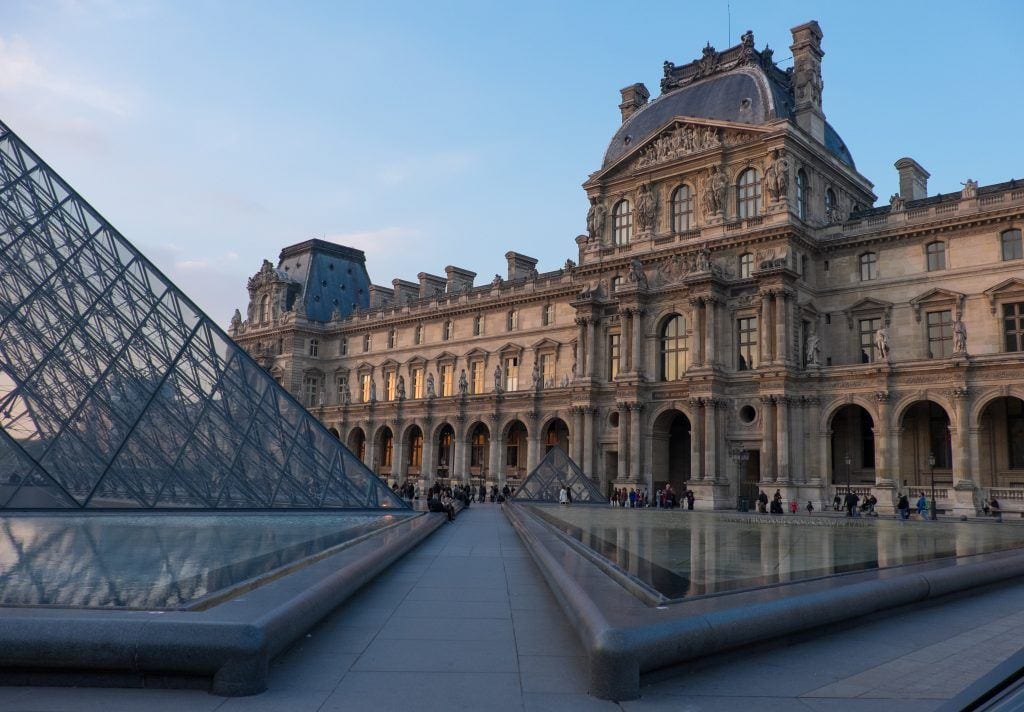 Exterior of the Louvre Museum in Paris with an ornate building covered in columns and arches, plus glass pyramids